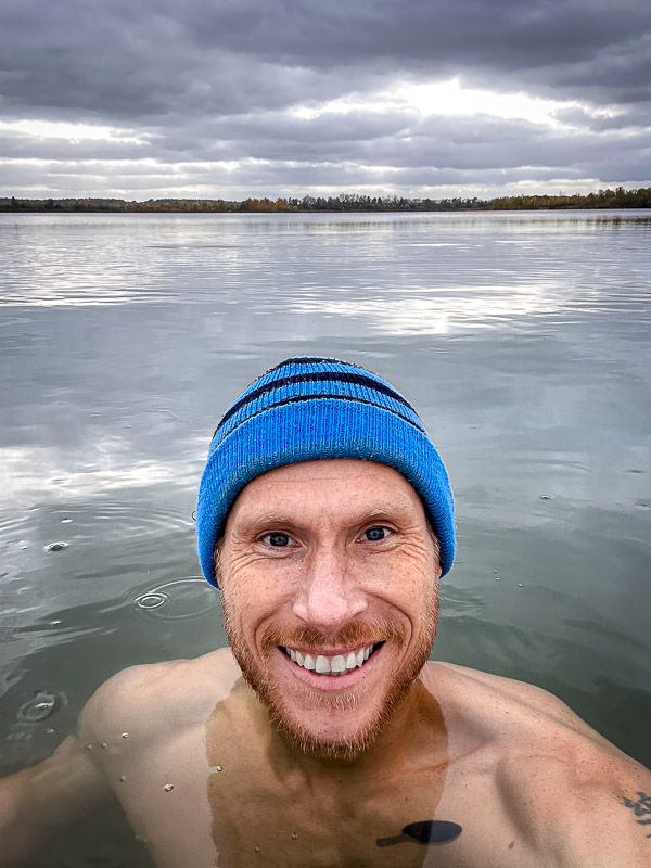 Een man met een blauwe muts lacht naar de camera terwijl hij in een koude meer staat. De lucht is bewolkt en het rustige water weerspiegelt de grijze wolken.