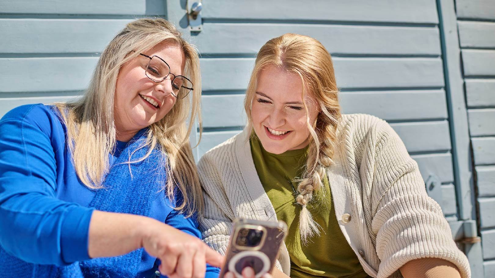 Twee vrouwen zitten buiten voor een blauwe houten wand en lachen, terwijl ze samen naar een smartphone kijken.