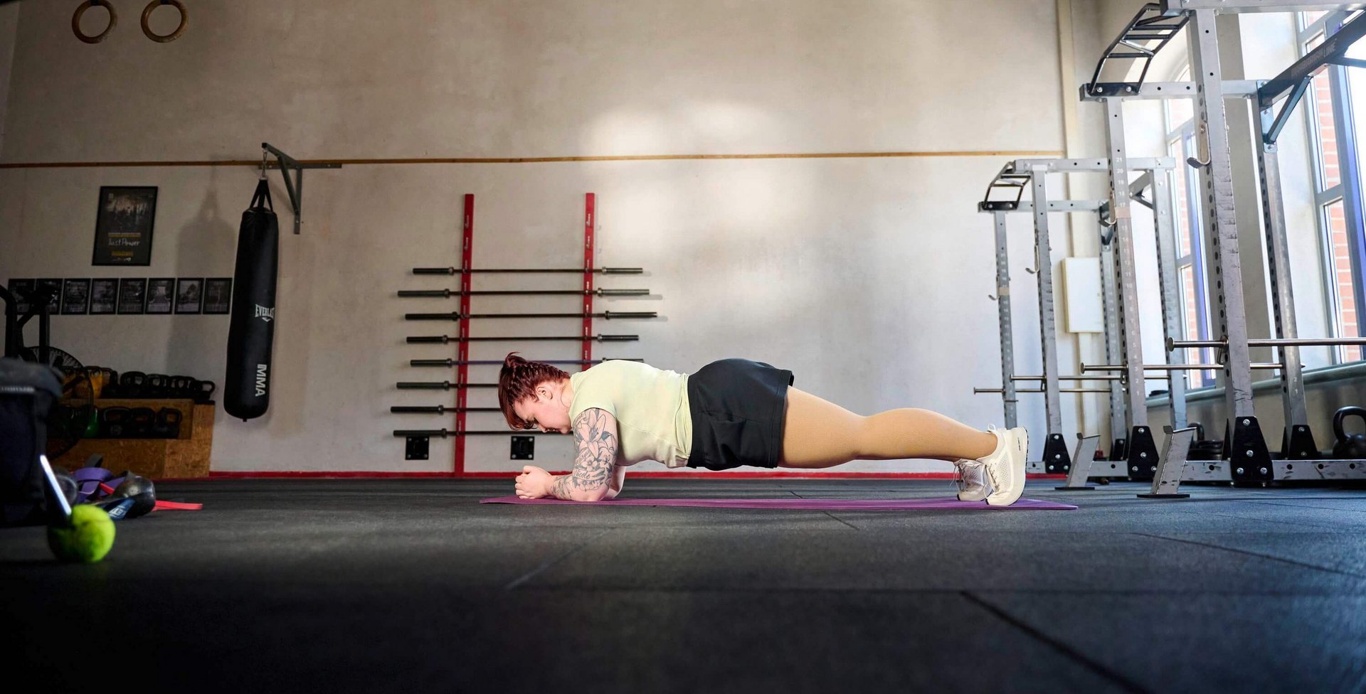 Persoon doet een onderarmsteun-oefening (plank) op een fitnessmat in een ruime trainingsruimte met hoge plafonds, halterstangen aan de wand en ramen aan de rechterkant.