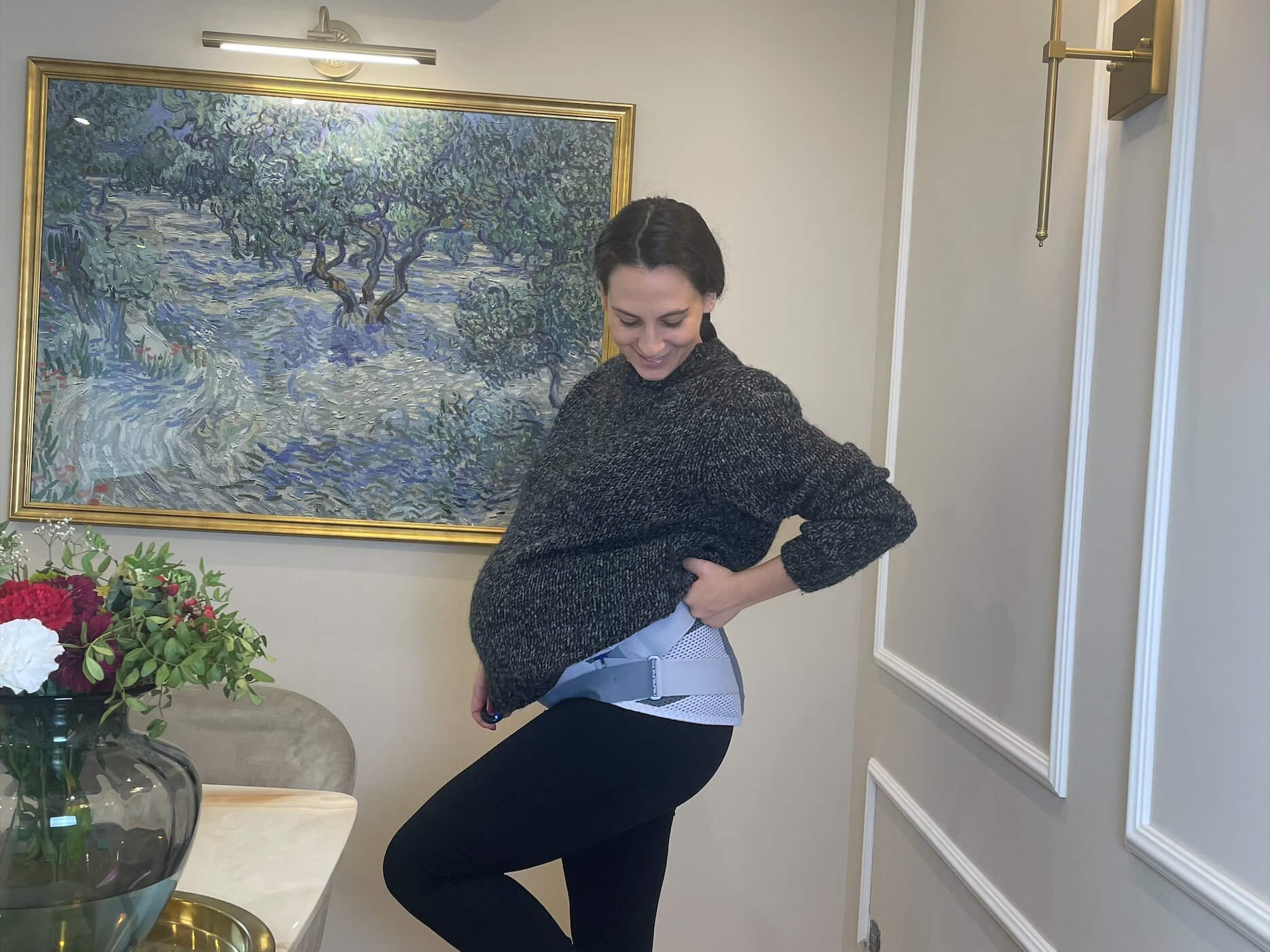 Zwangere vrouw staat op één been in een modern ingerichte ruimte, trekt haar donkere trui omhoog en toont een zwangerschapsorthese die over de zwarte legging zit.