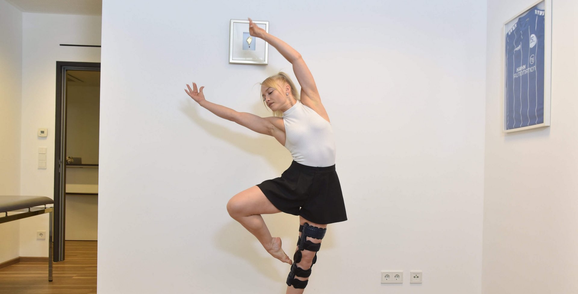 Anne-Maria Kot danst op blote voeten in een ruimte, draagt een zwarte rok, een wit mouwloos topje en een SecuTec Genu Flex knie-orthese aan haar linkerbeen.
