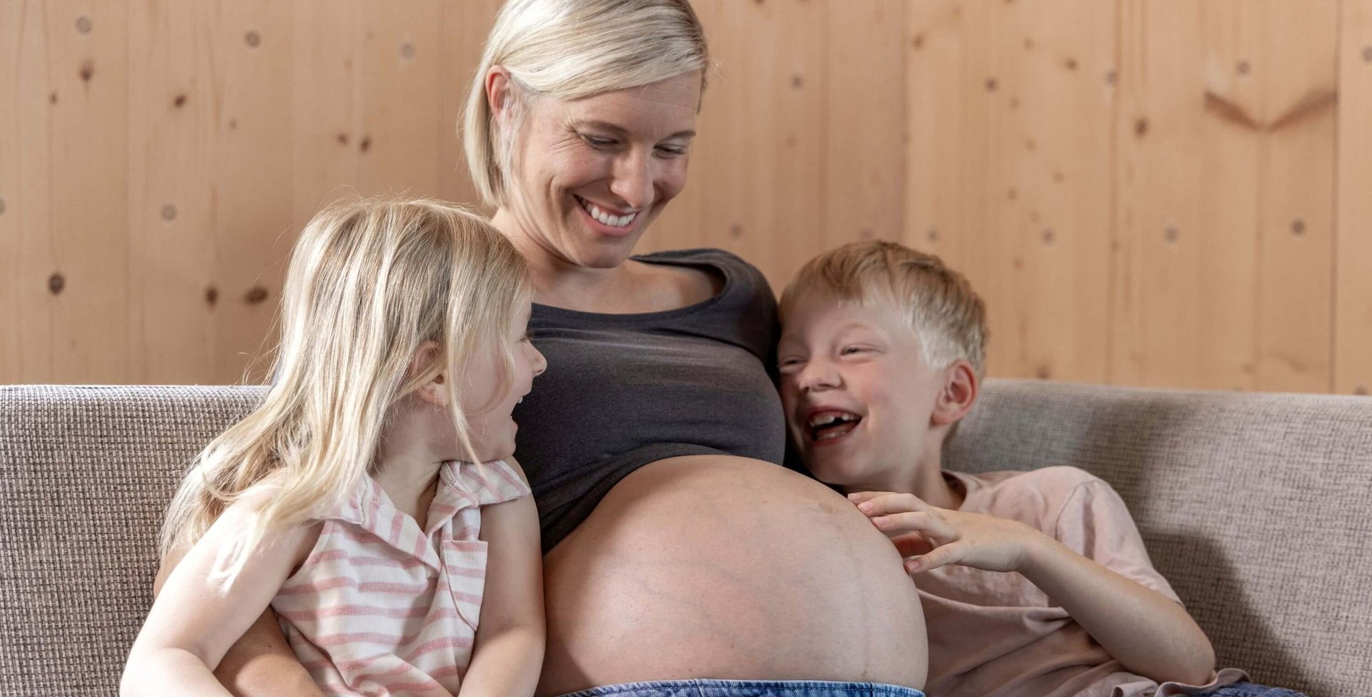 Zwangere vrouw zit op een bank en lacht, terwijl haar twee kinderen vrolijk haar babybuik aanraken.