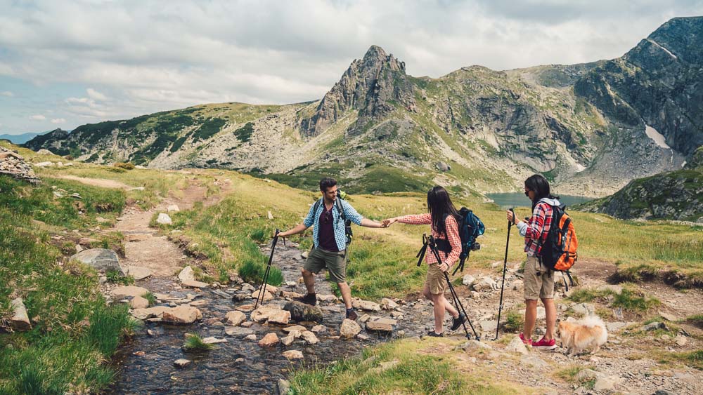 Drie wandelaars, twee vrouwen en een man, steken samen een bergbeek over in een alpenlandschap met rotsachtige toppen op de achtergrond. De man helpt een vrouw – de nadruk ligt op teamwork en natuurbeleving.