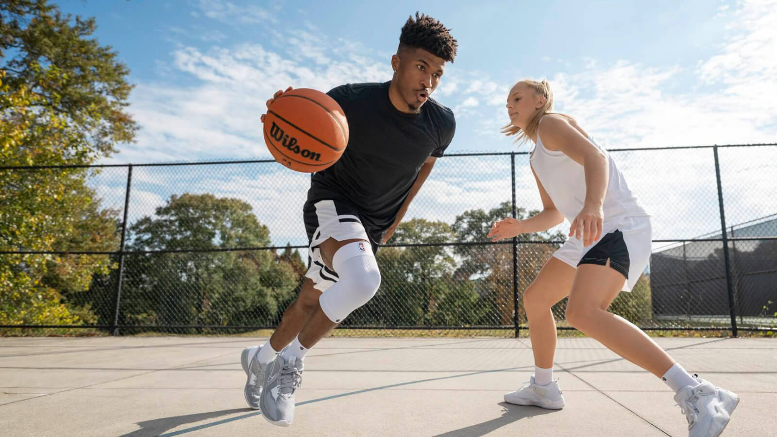 Een basketballer sprint over een buitenveld met de bal, op de hielen gezeten door een tegenstander. Hij draagt een witte kniebrace voor extra stabiliteit. De omgeving is herfstachtig met bomen en een hekwerk rondom.