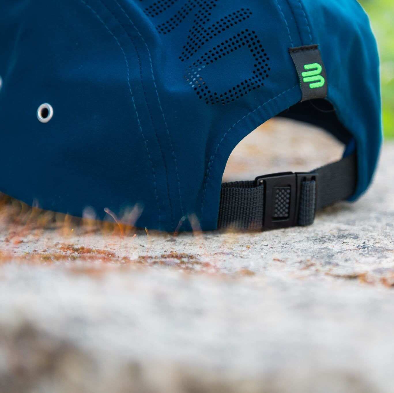 Close-up van de achterkant van een blauwe Bauerfeind Sports pet met zwarte verstelbare sluiting, groen logolabel, metalen oogje aan de zijkant en geperforeerde tekst – praktische details voor een stevige pasvorm tijdens actieve momenten buiten.