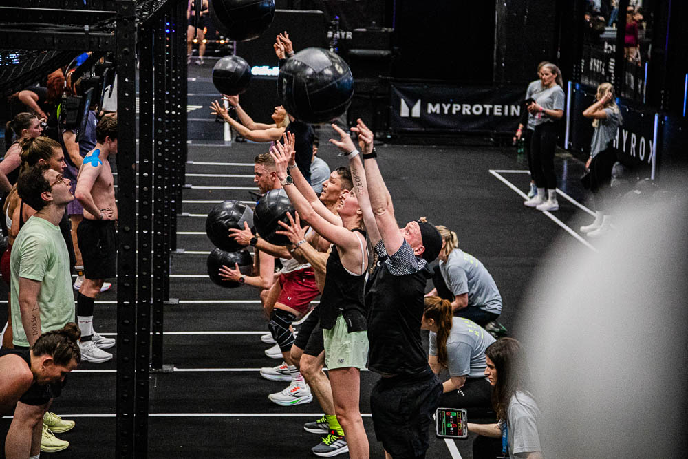 Atleten voeren wall balls uit in een HYROX-trainingszone en gooien zwarte medicine balls omhoog, terwijl juryleden en begeleiders de herhalingen volgen; de scène toont intensieve full-body inspanning, concentratie en een motiverende wedstrijdsfeer.