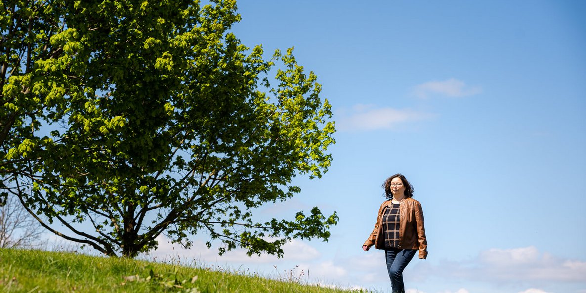 Katrin, een lymfoedeempatiënte, wandelt over een groene weide onder een heldere blauwe hemel. Op de achtergrond staat een grote volle boom die een zomerse en rustige sfeer uitstraalt. Ze draagt een bruin leren jasje en een spijkerbroek.