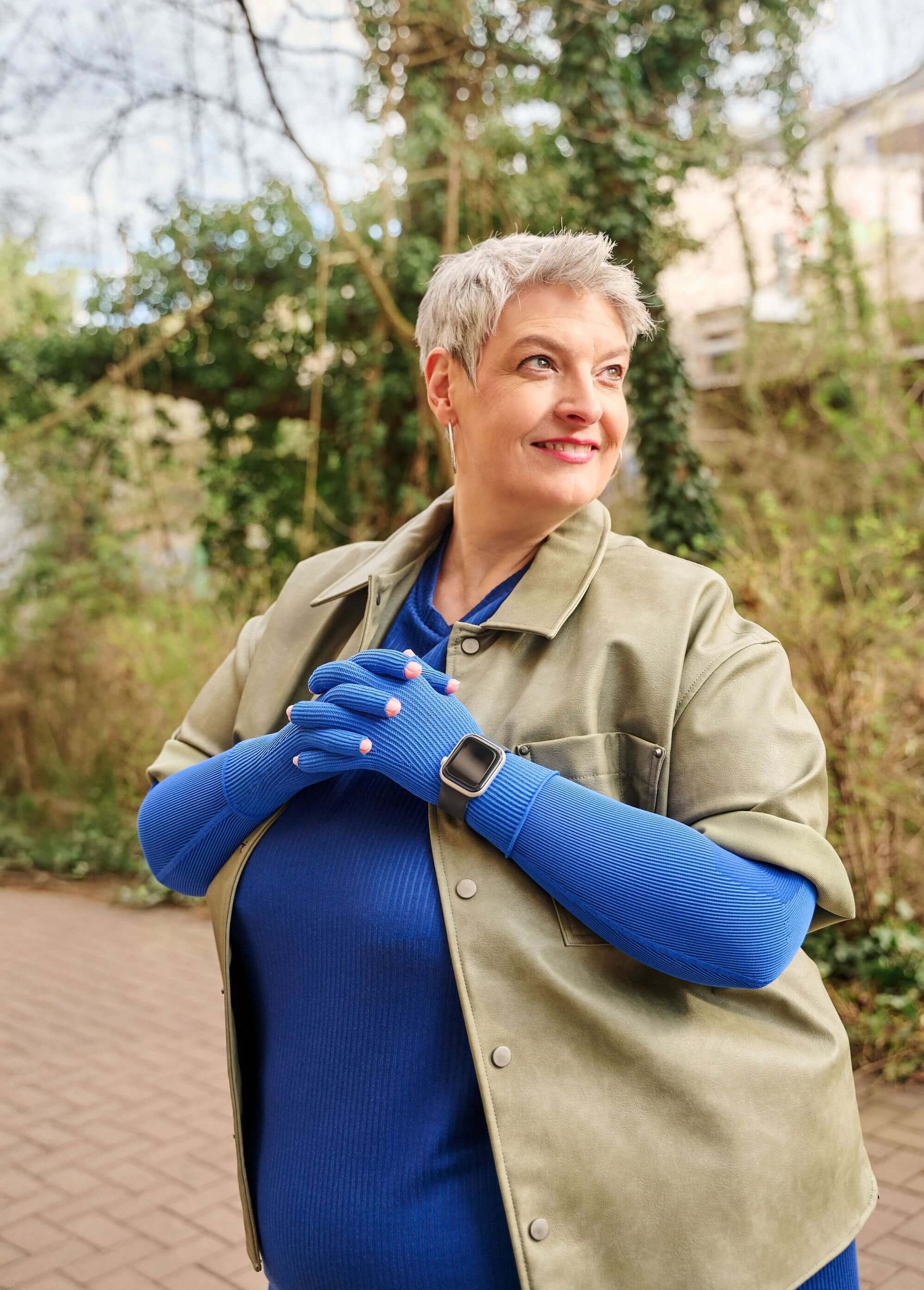 Oudere vrouw met kort grijs haar staat buiten op een geplaveid pad en lacht opzij. Ze draagt een blauw bovenstuk met bijpassende blauwe handschoenen, een olijfgroene jas en een smartwatch; op de achtergrond bomen en klimplanten.