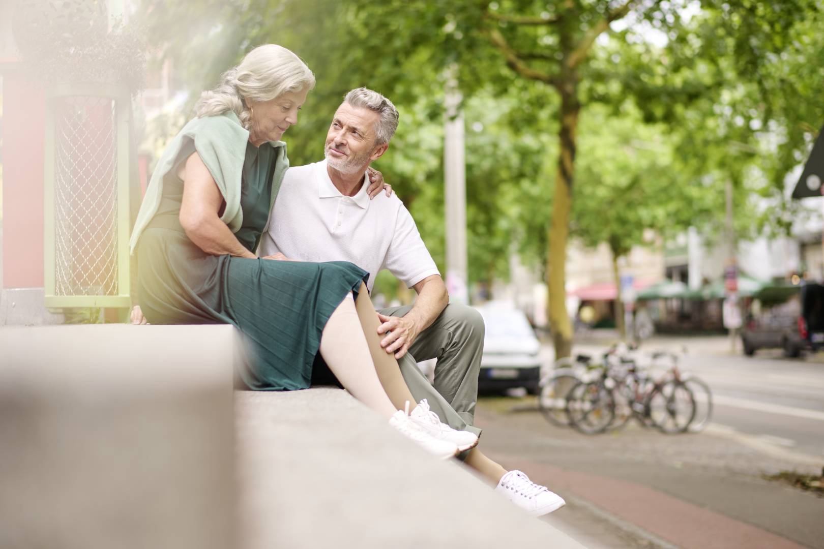 Een ouder stel zit ontspannen in een stedelijke omgeving. De vrouw draagt een groene jurk en compressiekousen uit de VenoTrain Special-lijn. De man legt liefdevol zijn hand op haar been.