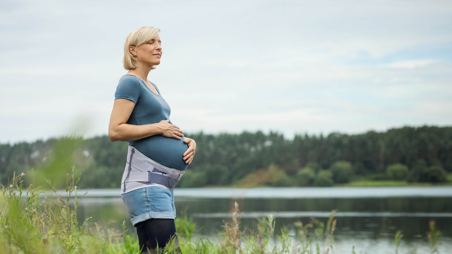 Zwangere vrouw staat aan de oever van een meer en houdt haar babybuik vast terwijl ze in de verte kijkt. Ze draagt de Spinova Mum zwangerschapsorthese en geniet van het rustige natuurdecor.