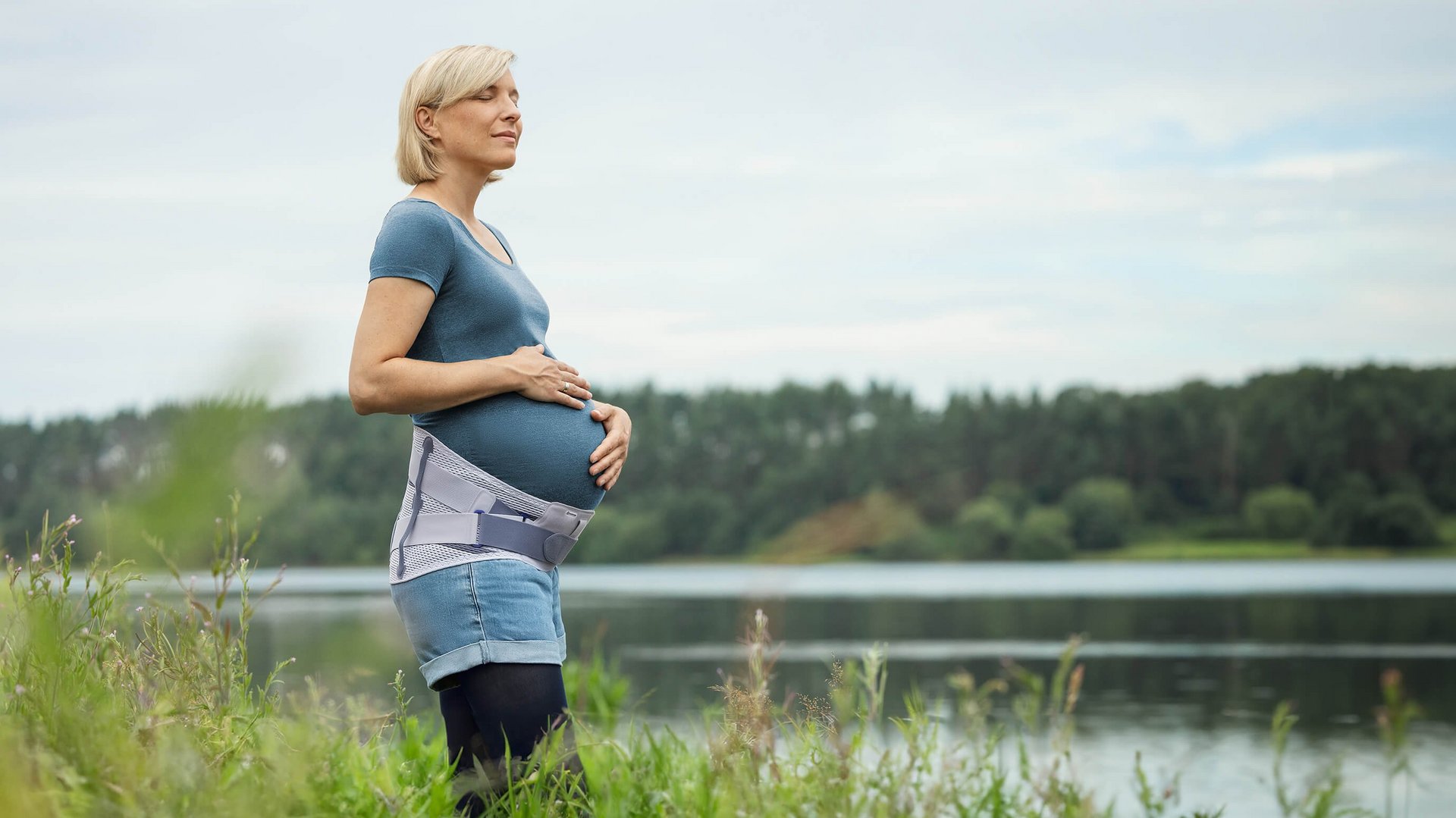 Zwangere vrouw staat aan de oever van een meer en houdt haar babybuik vast terwijl ze in de verte kijkt. Ze draagt de Spinova Mum zwangerschapsorthese en geniet van het rustige natuurlandschap.