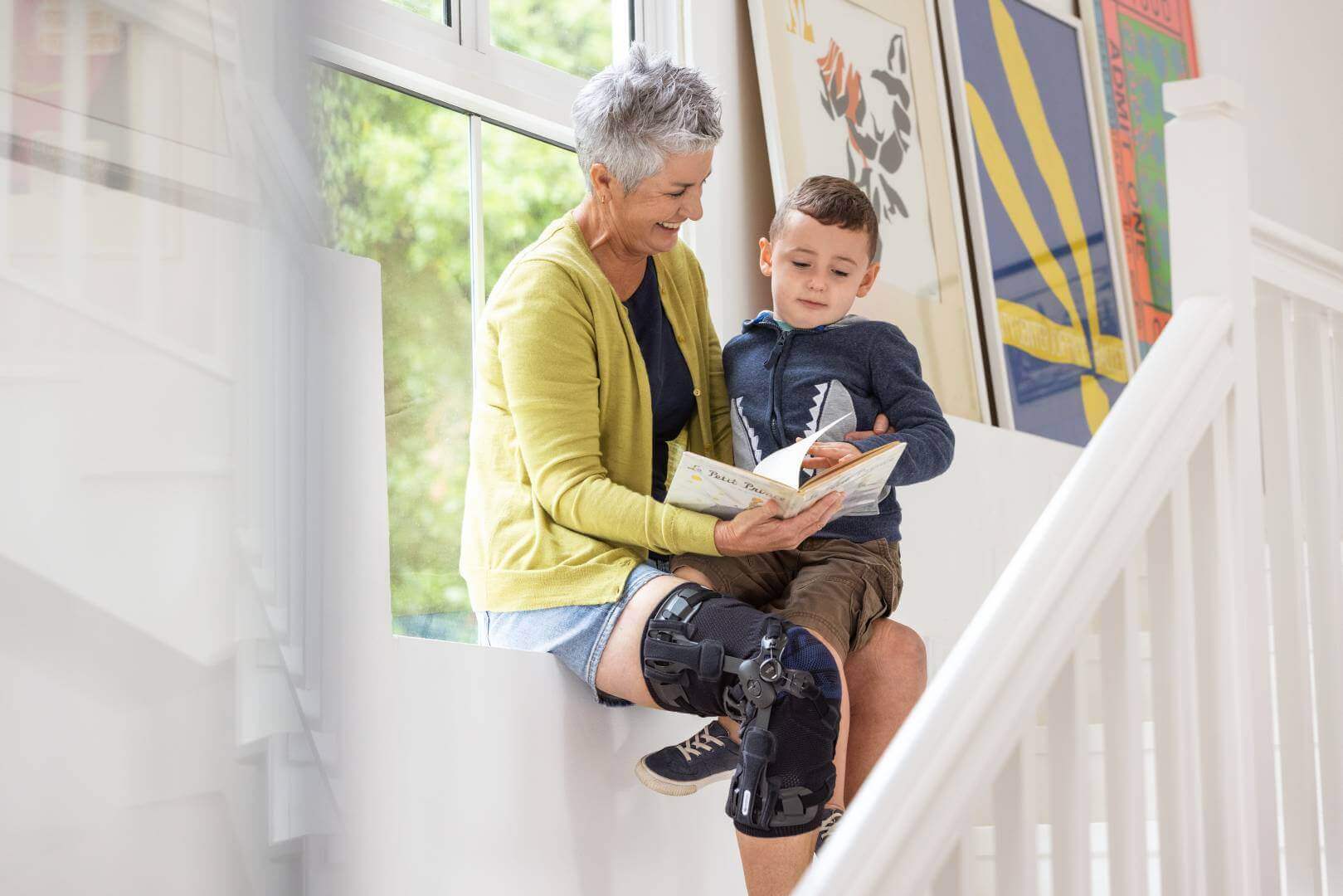 De afbeelding toont een oudere vrouw die samen met haar kleinzoon in een boek leest. De dame draagt een GenuTrain OA brace voor de conservatieve artrosebehandeling.