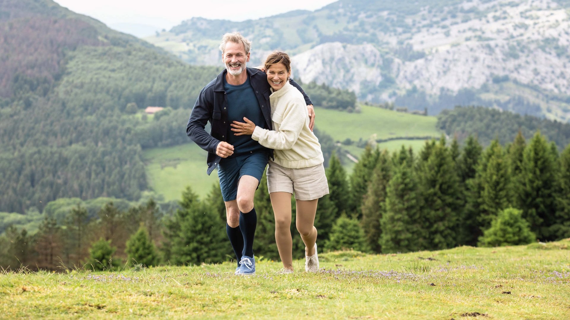 Een man en een vrouw lopen arm in arm in een berglandschap over een weiland. Beiden dragen VenoTrain impuls compressiekousen van Bauerfeind. Die helpen bij couperose.