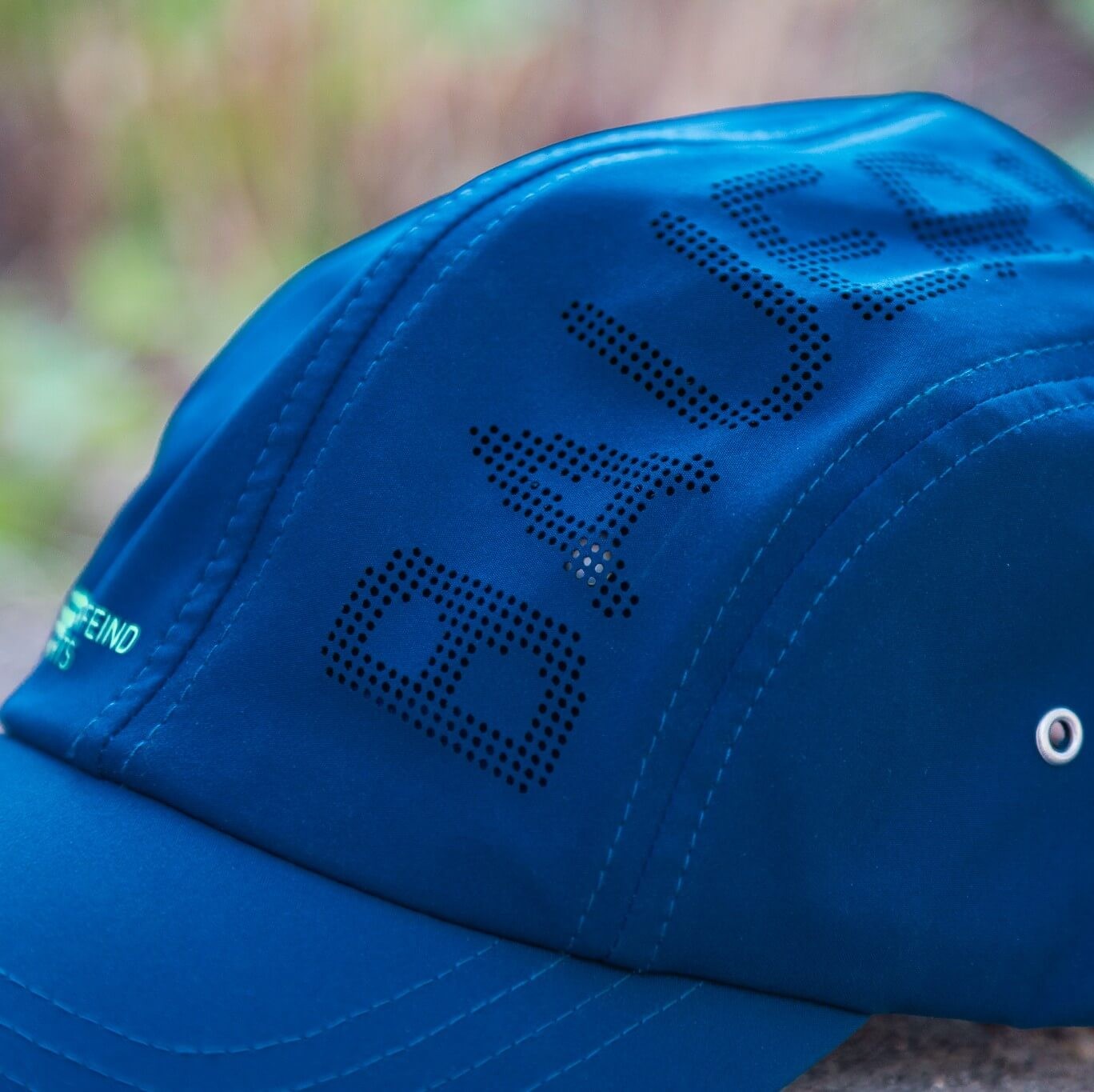 Close-up van de blauwe Bauerfeind Sports pet met geperforeerde BAUERFEIND-tekst, zichtbare stiksels, glad functioneel materiaal en een metalen oogje aan de zijkant – sportief design met focus op comfort en ventilatie.