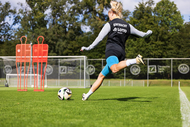 Een speelster van Eintracht Frankfurt schiet met volle kracht op het doel op de achtergrond. Ze draagt trainingskleding met het Eintracht-logo en een blauwe knieband van Bauerfeind Sports.