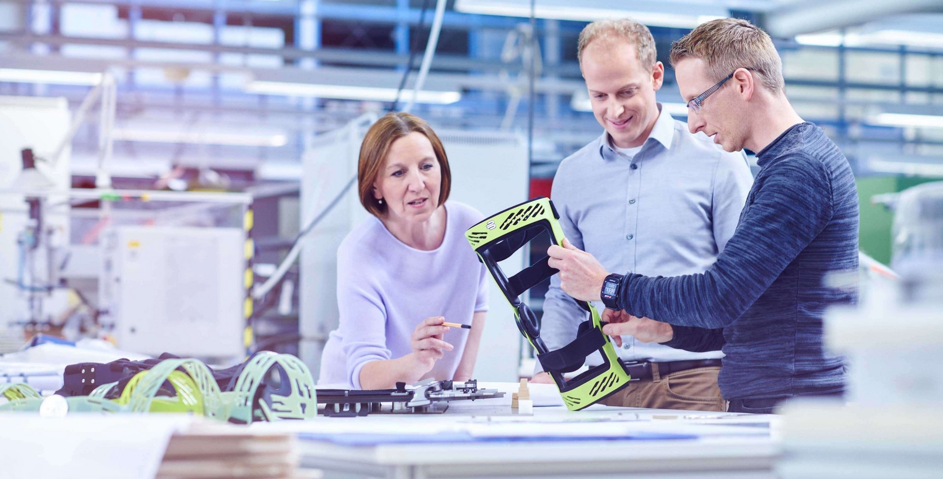 Drie personen in een werkhal bekijken een groene frame-orthese van Bauerfeind, één persoon houdt hem omhoog, op de tafel liggen technische tekeningen en gereedschap.