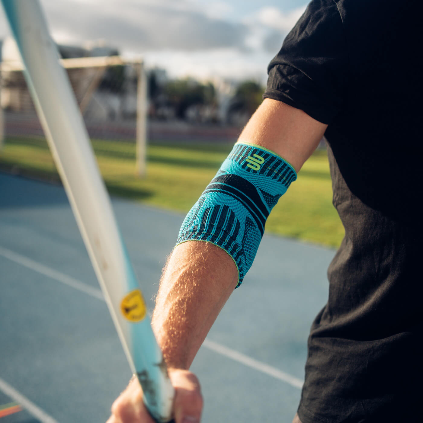Close-up van een arm met een blauwe elleboogbrace tijdens het speerwerpen. Op de achtergrond zijn een atletiekbaan en een zonnig landschap te zien.