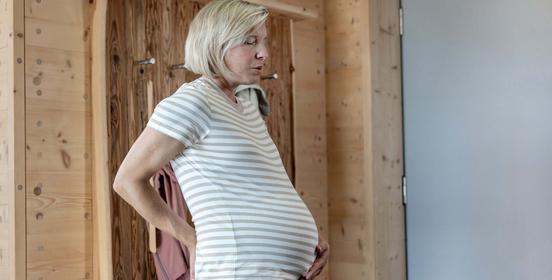 De afbeelding toont een zwangere vrouw die haar handen tegen haar onderrug steunt en diep ademhaalt. De scène toont typische rugpijn tijdens de zwangerschap.
