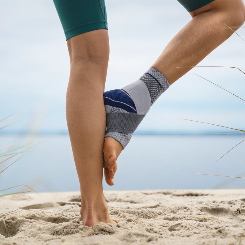 Close-up van persoon die stretcht op strand met grijs-blauwe MalleoTrain Plus enkelbrace.