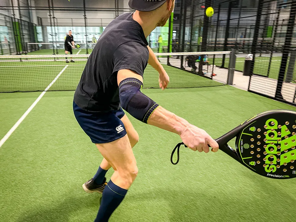 Een padelspeler in een zwart shirt en korte broek slaat de bal met volle concentratie. Hij draagt een donkerblauwe sportbrace om zijn rechterarm voor extra ondersteuning.