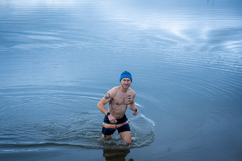 Een man waadt uit het meer, gefilmd tijdens een ijsbad in zwembroek en blauwe muts in de herfstomgeving.