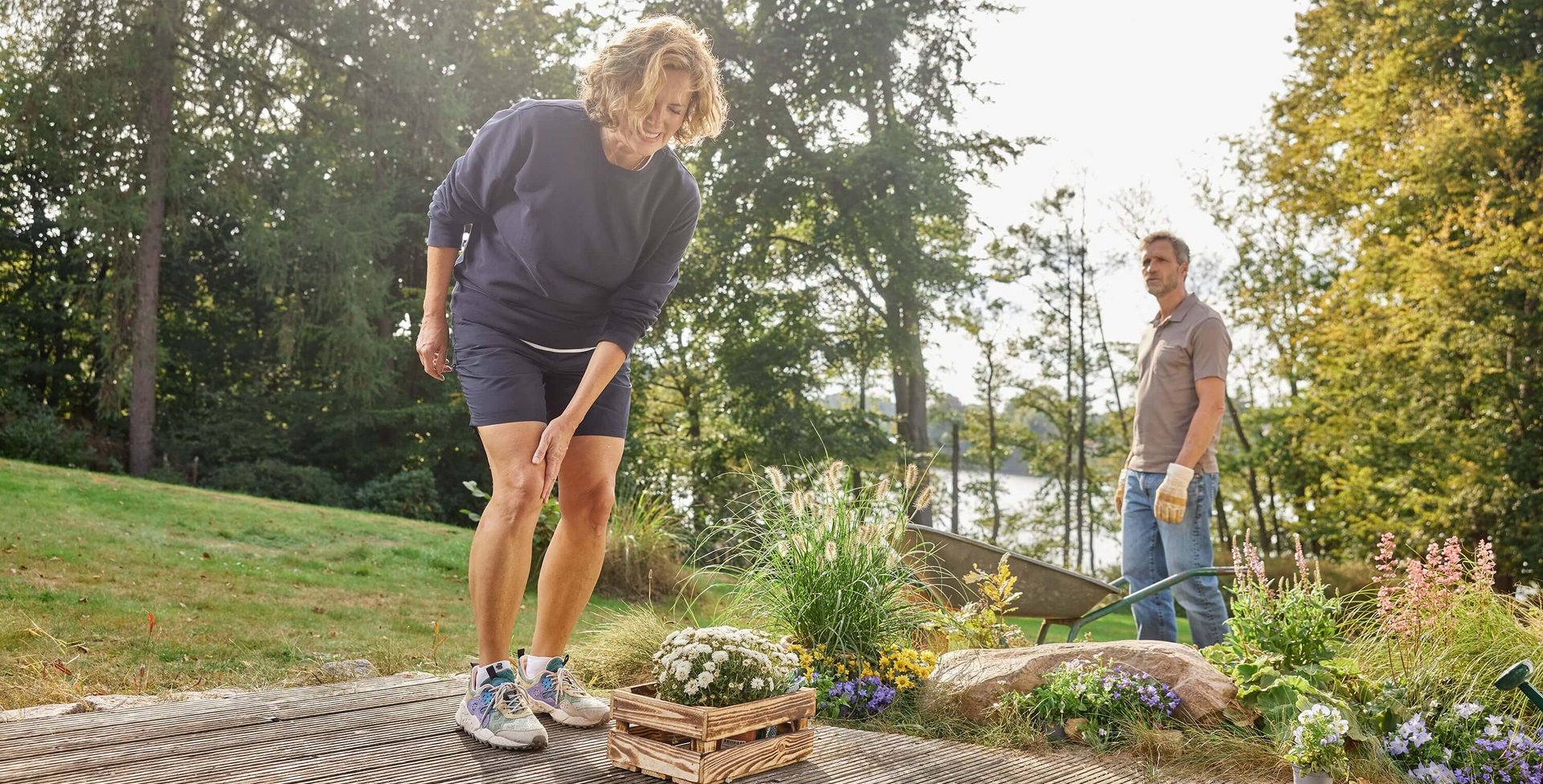 Een vrouw grijpt tijdens het tuinieren naar haar pijnlijke knie. Op de achtergrond staat een man met handschoenen toe te kijken. De tuin is omgeven door kleurrijke bloemen en een idyllische omgeving met bomen en een meer.