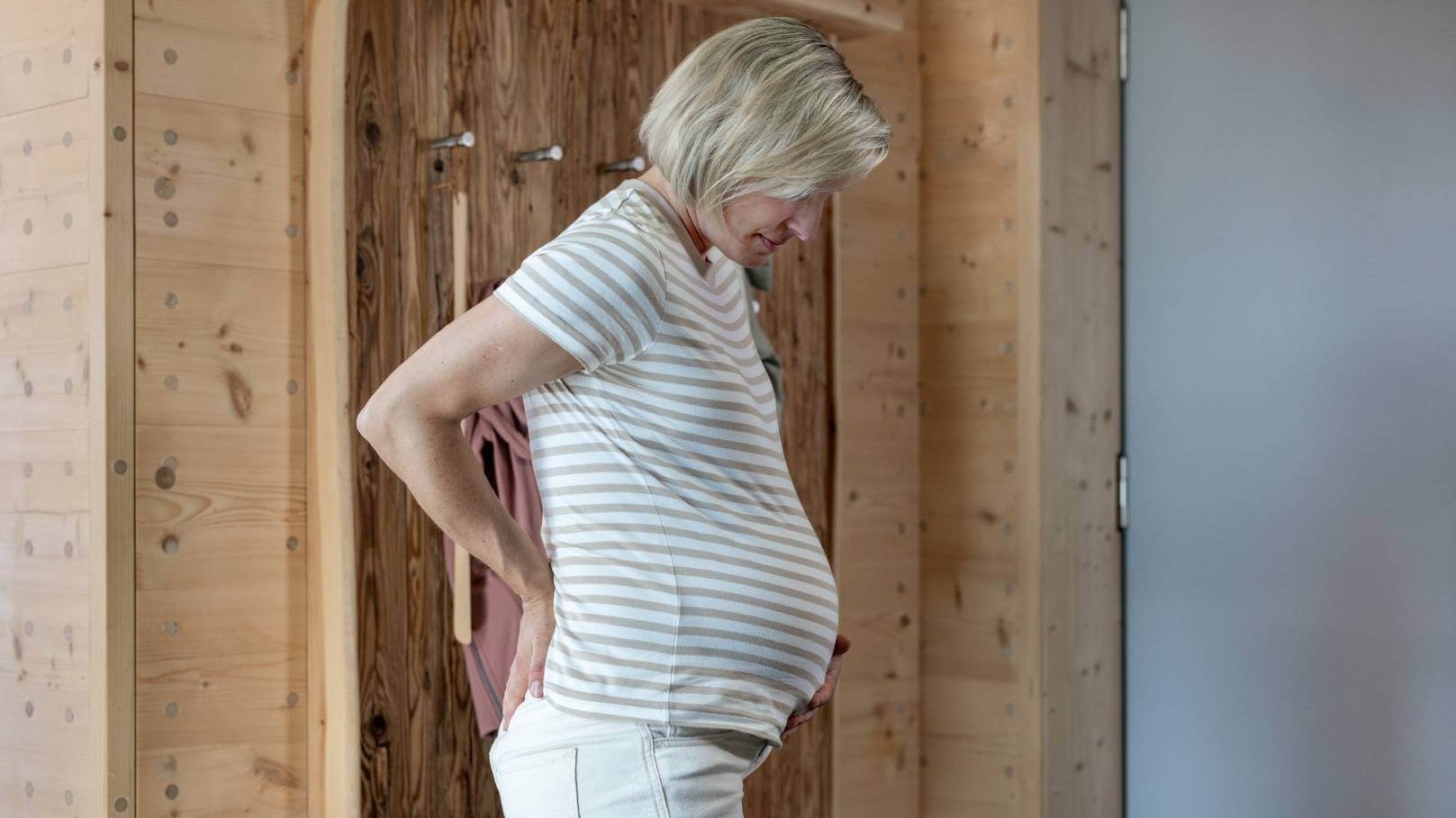 Een zwangere vrouw staat in een ruimte met houten wanden en steunt haar handen tegen haar pijnlijke onderrug terwijl ze naar haar babybuik kijkt.