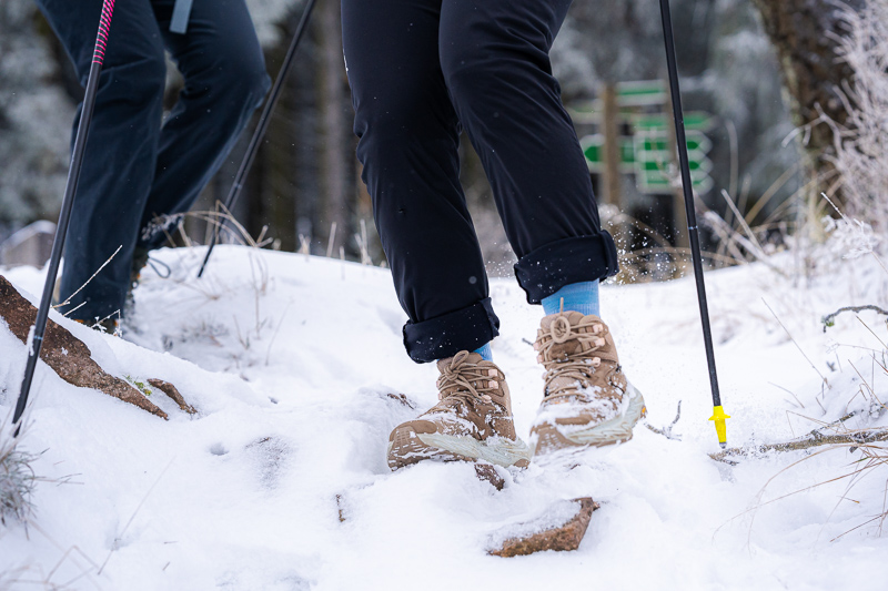 Close-up van wandelschoenen die door diepe sneeuw stappen. De voeten behoren toe aan wandelaars met zwarte broeken en blauwe sokken. Wandelstokken met gele punten staan in de sneeuw, terwijl de grond bedekt is met sneeuw en stenen.