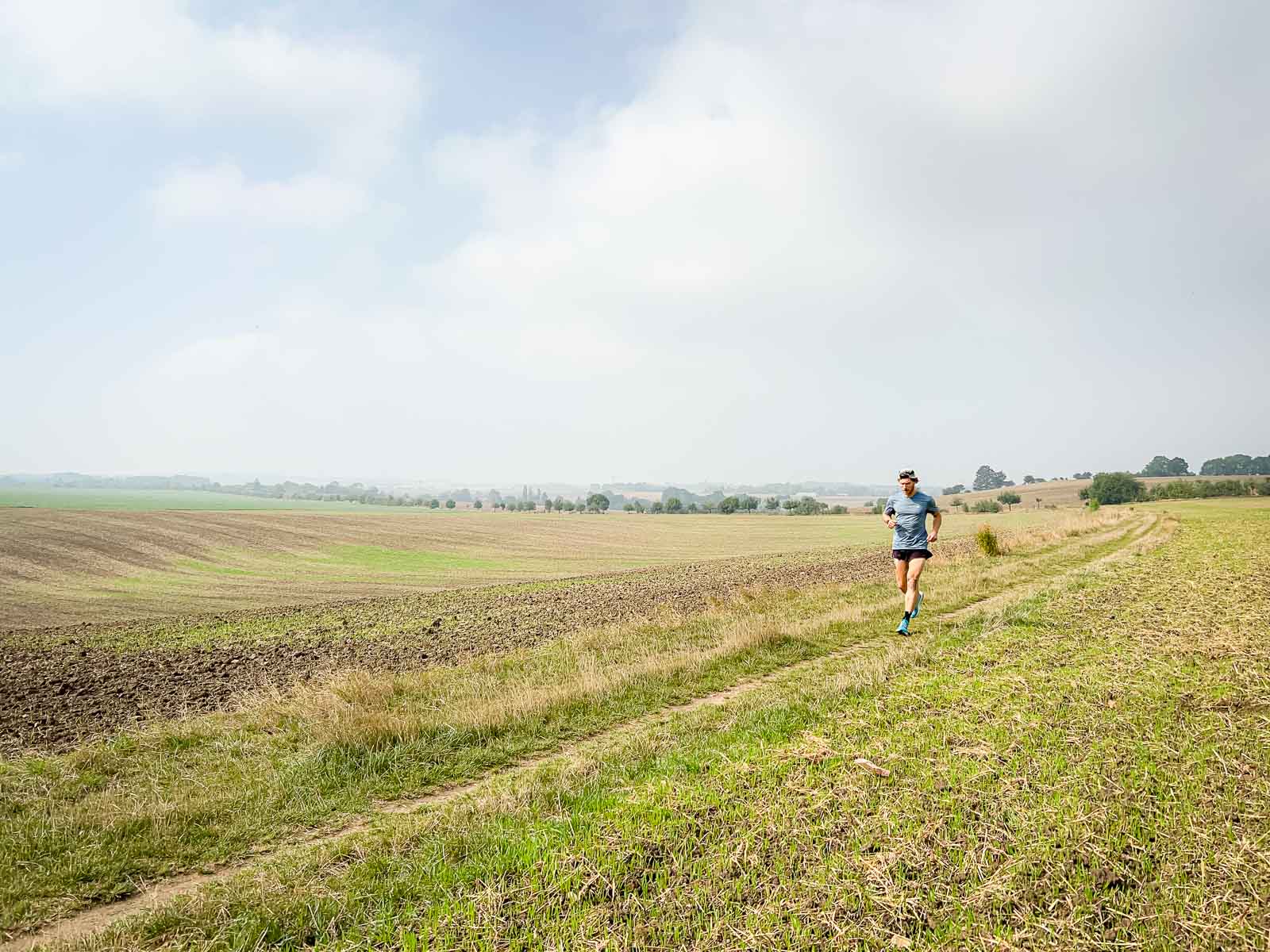 Een loper rent op een veldweg in een landelijke omgeving met een lichtbewolkte hemel. De omgeving is rustig en uitgestrekt, perfect voor duurtraining.