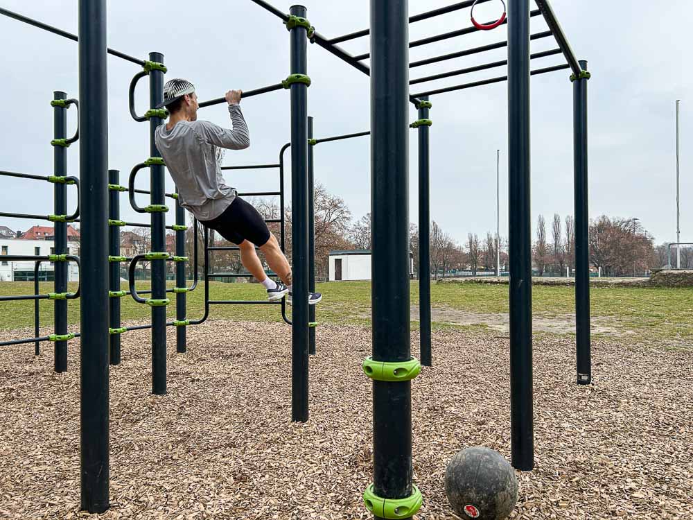 Atleet doet tijdens HYROX-training aan een outdoor calisthenicsrek een trekoefening aan de stang; op de grond ligt een zwarte trainingsbal.