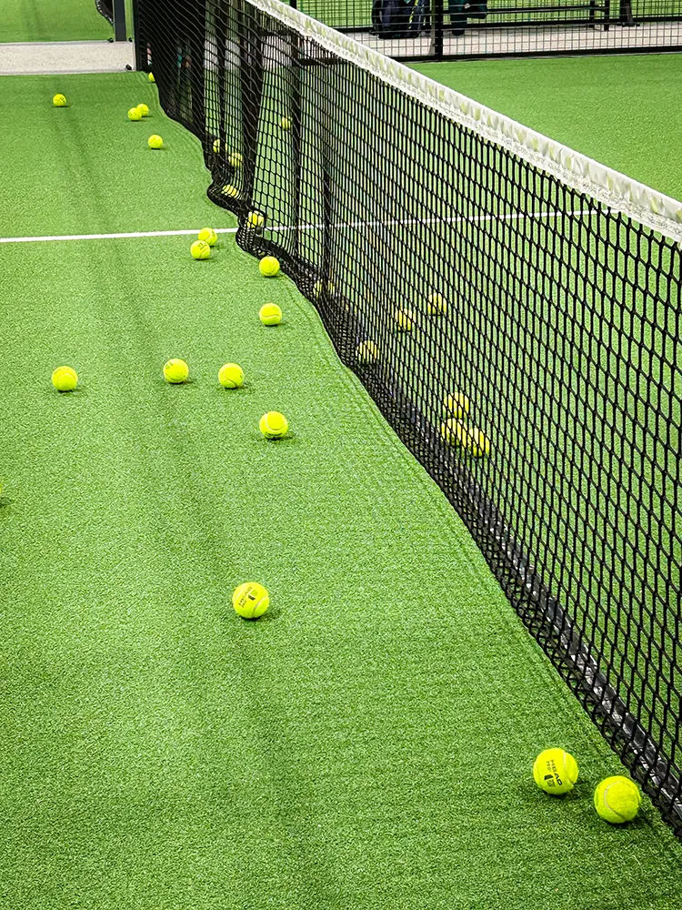 Meerdere gele padelballen liggen verspreid over het groene kunstgras naast het net – een levendig beeld na intensieve rally's.