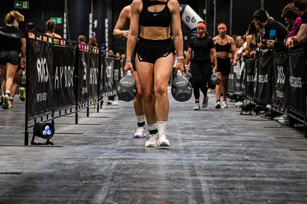 Atlete draagt tijdens een HYROX-wedstrijd twee zware kettlebells over het parcours, terwijl andere deelnemers achter haar volgen en toeschouwers langs de baan staan; de scène laat gripkracht