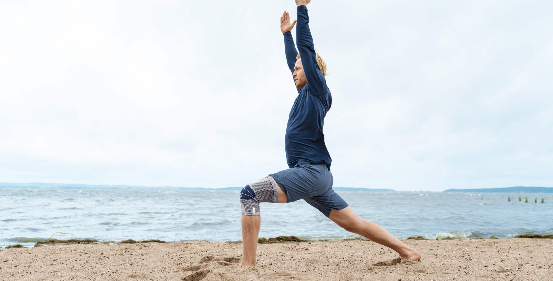 De afbeelding toont een man in yogahouding op het strand. Aan zijn rechterknie draagt hij een GenuTrain ter ondersteuning. De bandage biedt onder andere ondersteuning bij een vochtophoping in de knie.