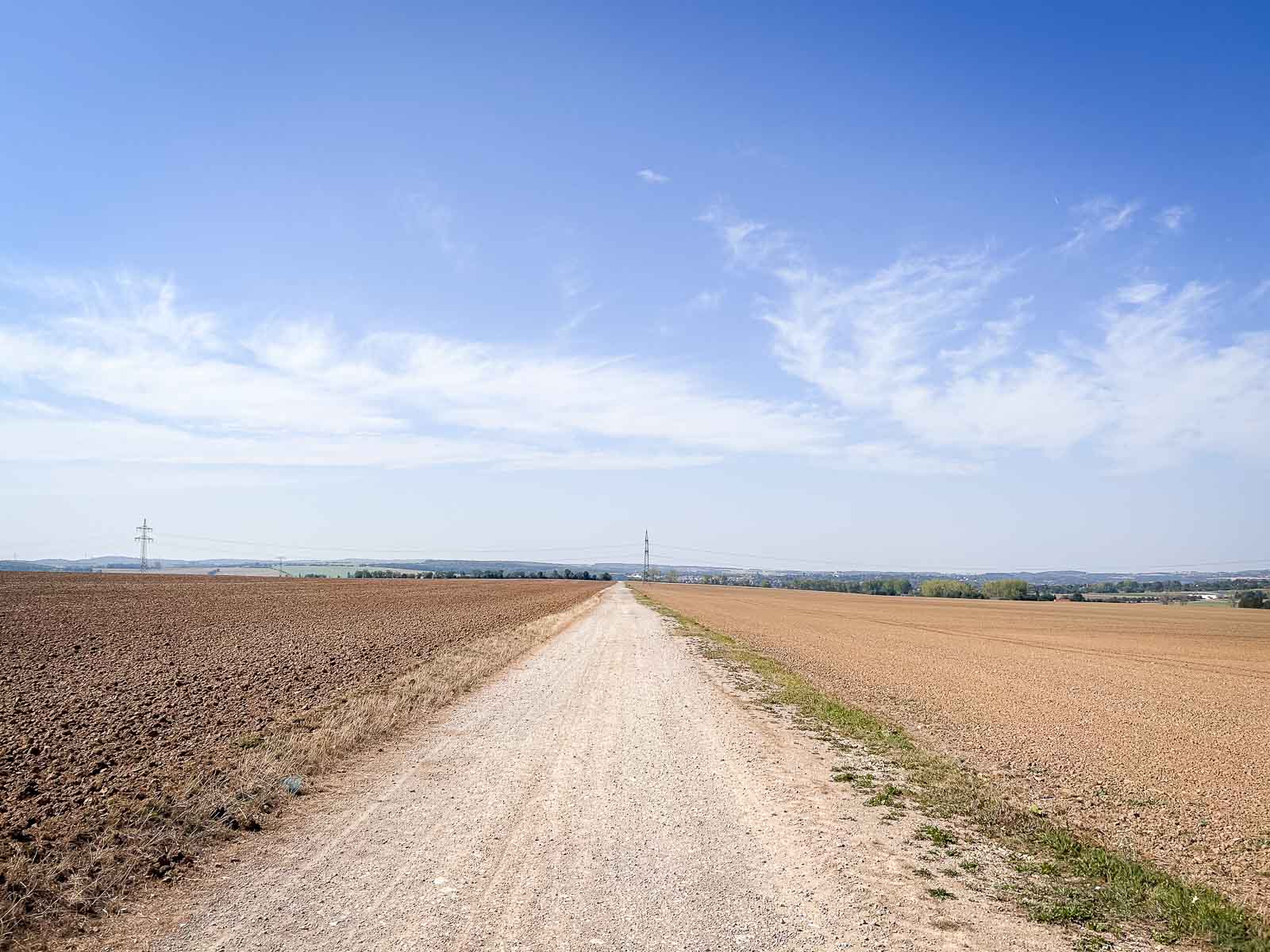 Een lange, rechte grindweg die door een uitgestrekt landschap loopt, met een heldere lucht en velden aan beide zijden. Ideaal hardlooptraject voor duurlopen.