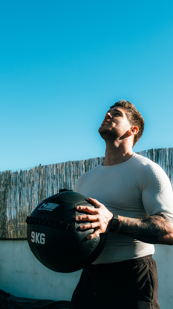Atleet staat buiten met een zwarte medicinebal van 9 kg voor het bovenlichaam, draagt een licht compressieshirt en kijkt omhoog onder een heldere blauwe lucht – functionele training voor hybrid athletes.