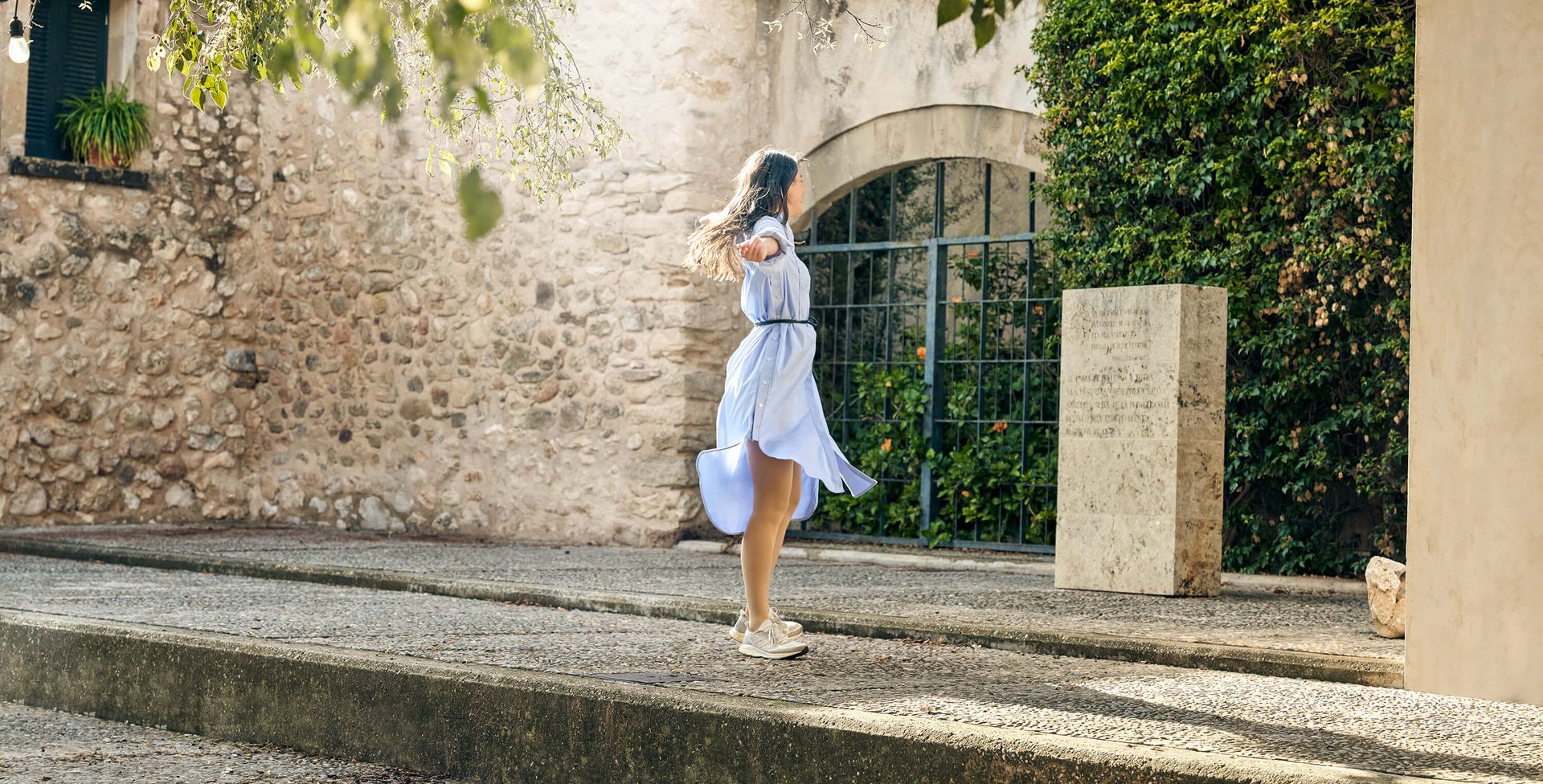 Een jonge vrouw in een zwierige lichtblauwe jurk draait met gespreide armen rond op een stenen terras voor een mediterrane stenen muur. Ze draagt ingetogen VenoTrain compressiekousen die harmonieus bij het outfit passen.