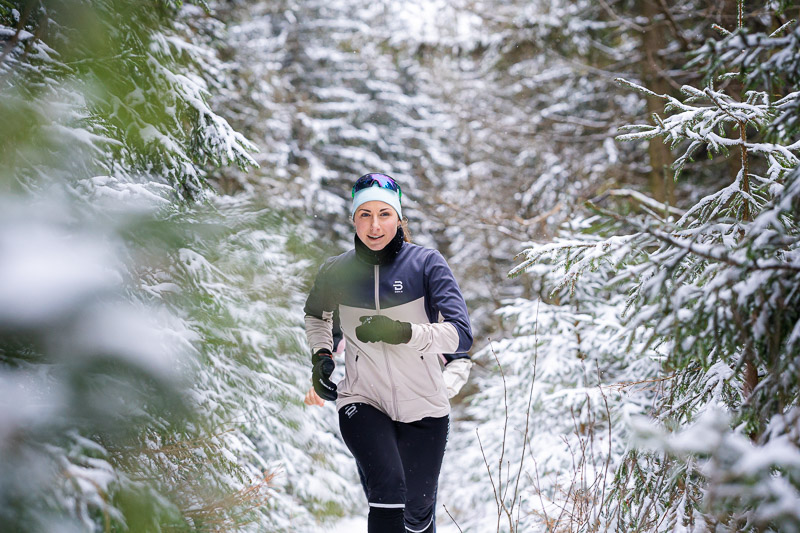Een hardloopster rent door een besneeuwd bospaadje. De bomen om haar heen zijn zwaar bedekt met sneeuw. Ze draagt een lichtgrijze hardloopjas met zwarte accenten, een zwarte broek en een blauwe hoofdband met een zonnebril.