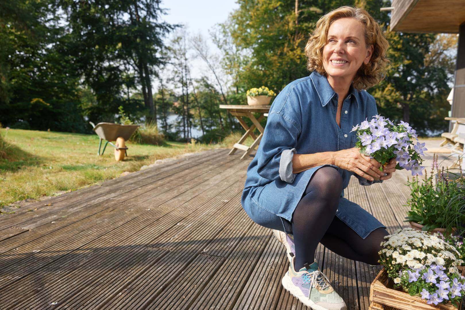 Een lachende vrouw in een spijkerjurk knielt op een houten terras en houdt een pot met paarse bloemen vast. Ze draagt VenoTrain soft compressiekousen en kleurrijke sportschoenen.