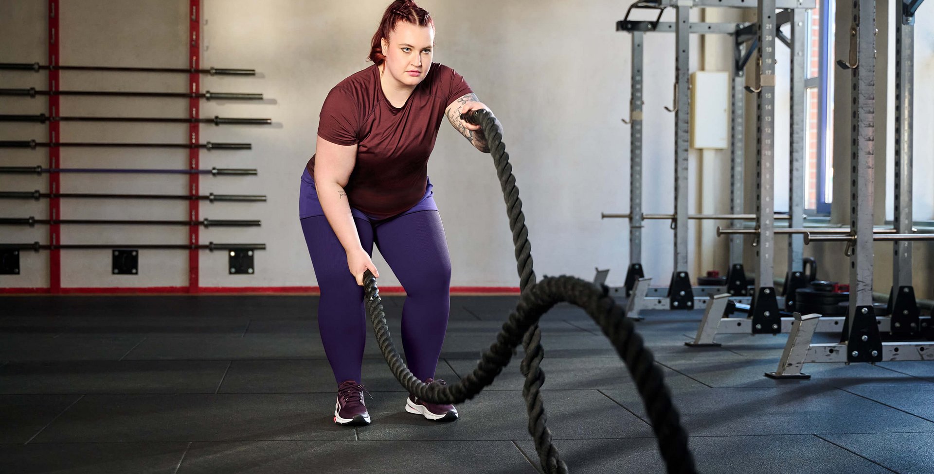 Een vrouw traint in een sportschool met battle ropes. Ze draagt een bordeauxrood T-shirt, paarse legging en sportschoenen. Op de achtergrond zijn halters aan de wand en krachttrainingstoestellen te zien.