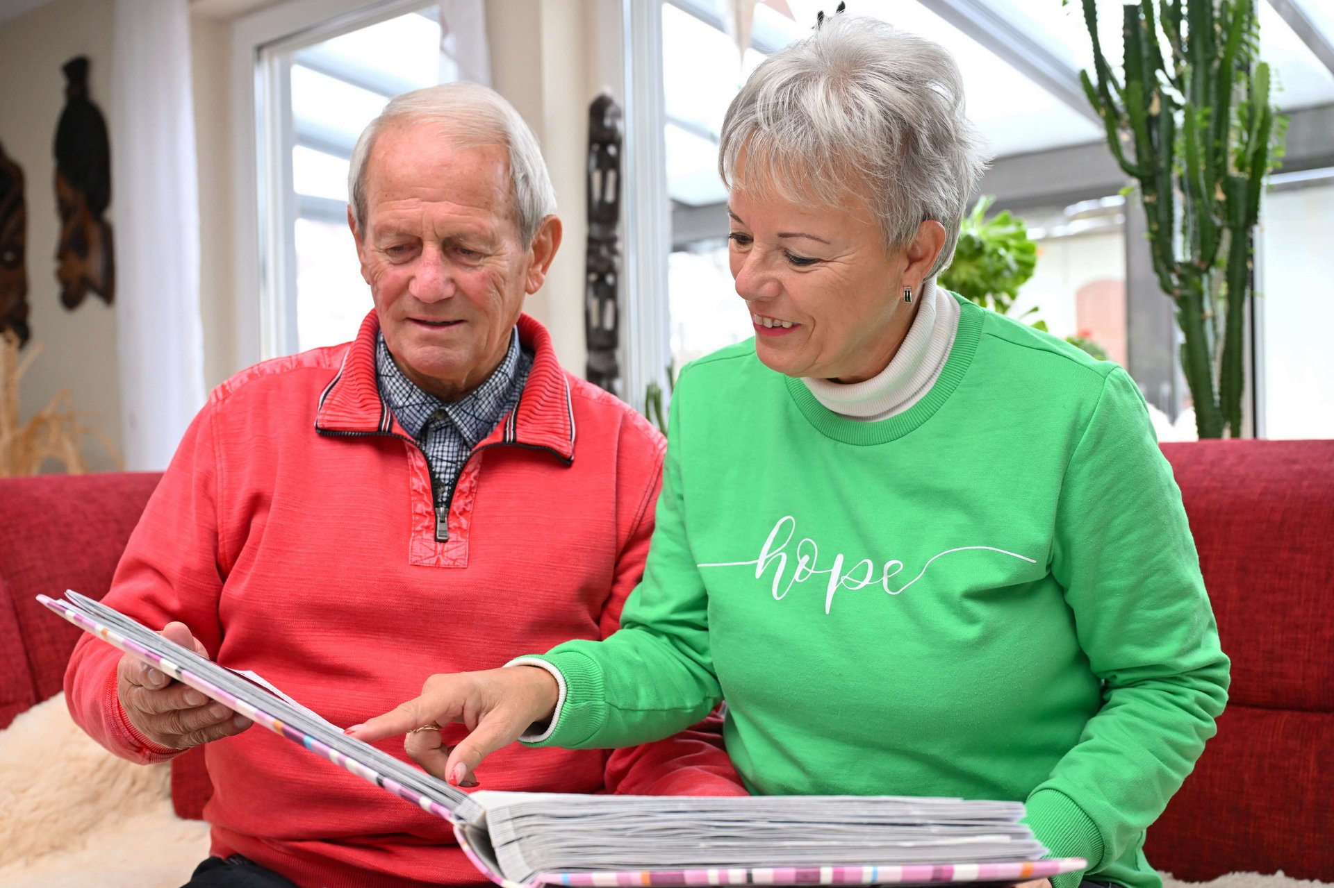 Lothar Schiller zit naast zijn vrouw op een rode bank in een lichte woonkamer. Samen bekijken ze een fotoalbum terwijl ze lachen en over de foto's praten.