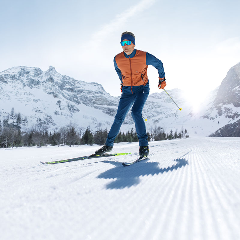 Een man met een blauwe skibril, van voren gefotografeerd, is aan het skiën in een winters landschap. Hij draagt een oranje jas en een blauwe skibroek.
