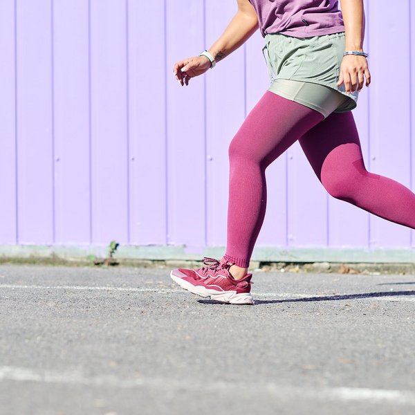 Een vrouw in sportieve kleding met een bordeauxrode panty en bijpassende sportschoenen loopt over asfalt voor een paarse houten wand. Alleen het onderlichaam en een deel van het bovenlichaam zijn zichtbaar.