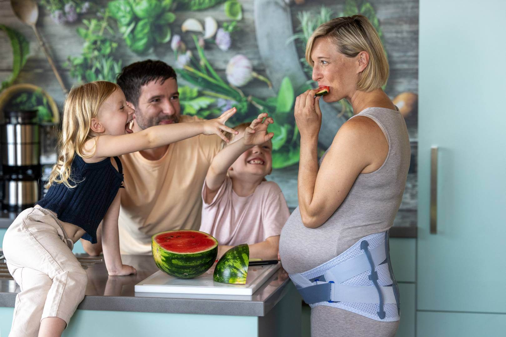 Een zwangere vrouw met Spinova Mum orthese eet een stuk watermeloen in de keuken, terwijl haar dochter lachend naar een stuk grijpt. De vader en de zoon kijken vrolijk toe.