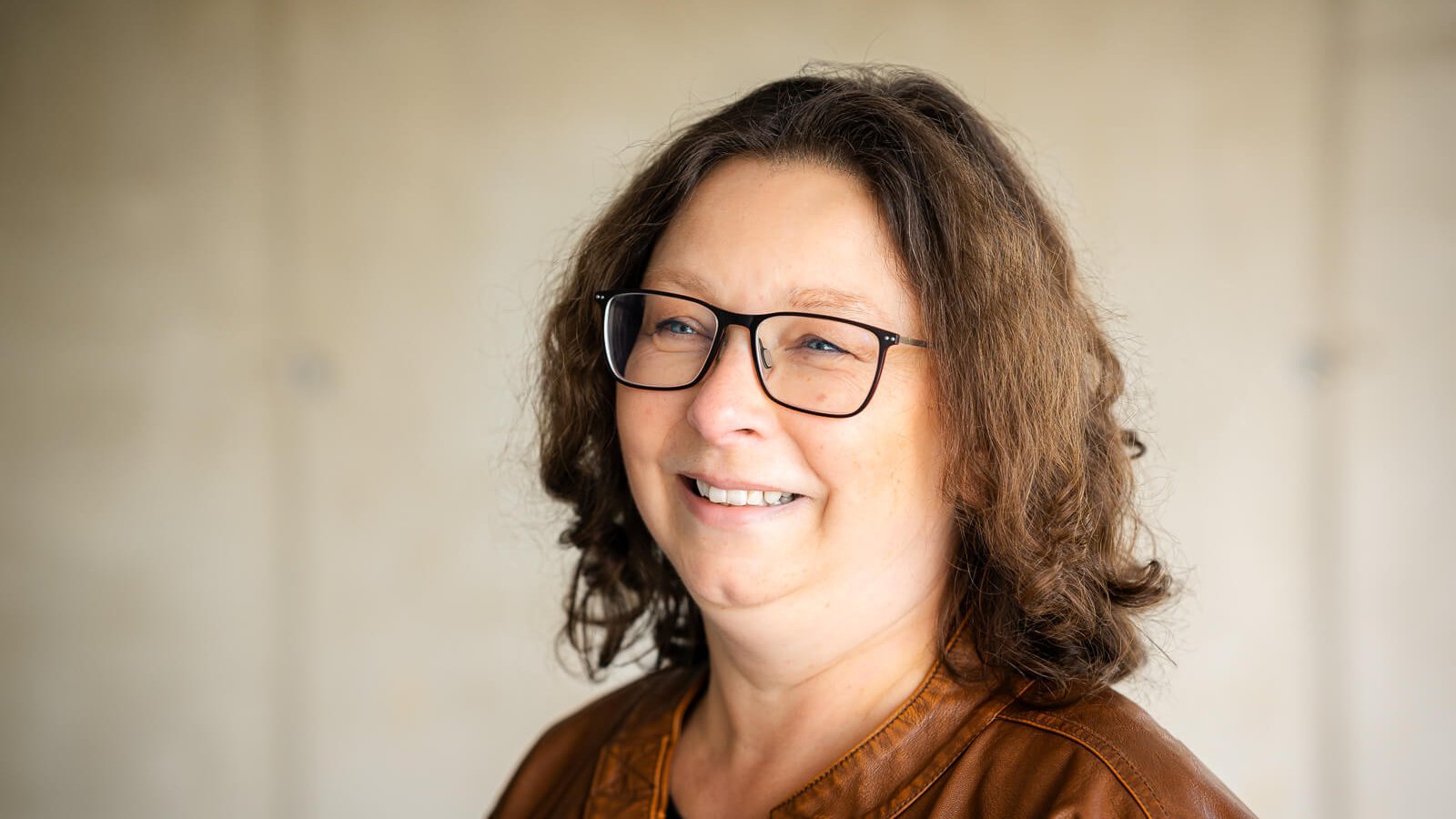 Portretfoto van Katrin, een lymfoedeempatiënte. Ze draagt een bril en een bruin leren jack. Op de achtergrond is een neutrale, onscherpe muur te zien. Katrin lacht vriendelijk opzij.