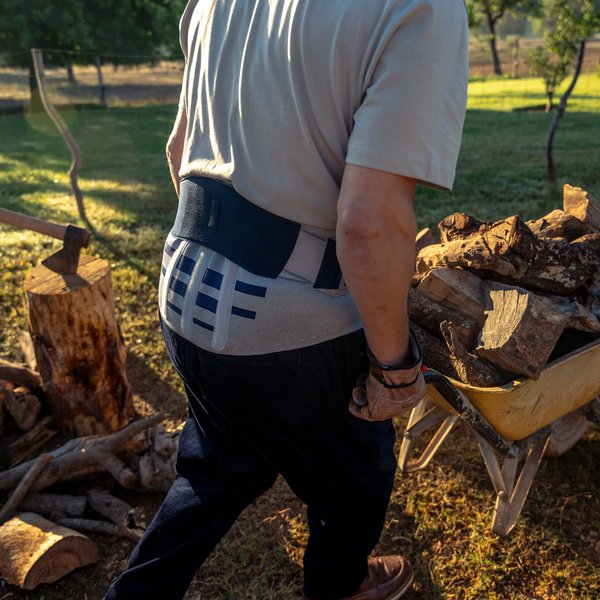 Een man met rugbandage van achteren, terwijl hij een gele kruiwagen vol houtstukken in de tuin verplaatst. Op de achtergrond staat een boomstronk met een bijl, omringd door houtblokken.