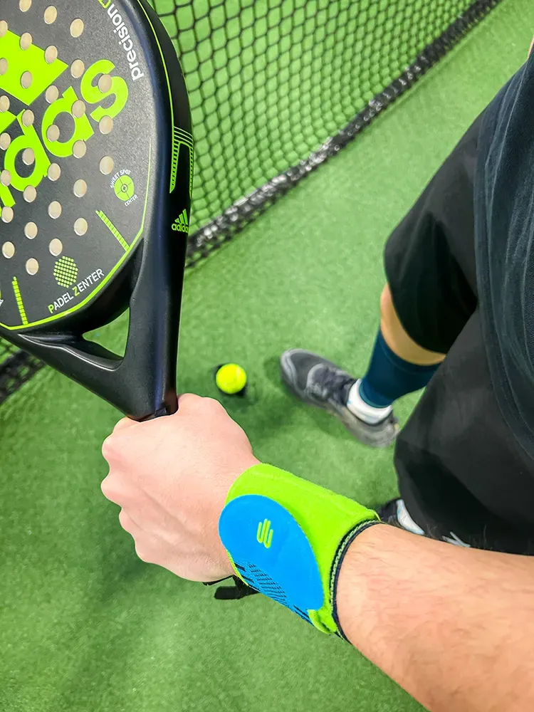 Nahaufnahme eines Spielers auf einem Padelplatz, der einen Adidas-Padelschläger hält. Der Sportler trägt eine neonfarbene Handgelenkbandage, während ein Ball am Boden liegt.
