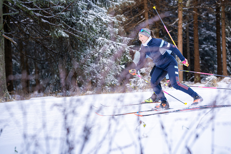 Een atlete die door een besneeuwd bos skiet. Ze beweegt energiek door diepe sneeuw, uitgerust met professionele ski-uitrusting en moderne sportkleding.