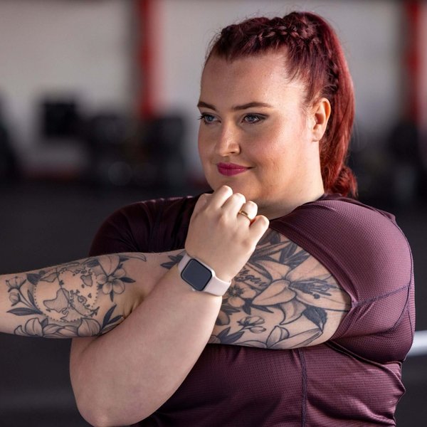 Vrouw met rood gevlochten haar en zichtbare tatoeages op beide armen rekt haar arm uit in een fitnessstudio. Ze draagt een bordeauxrood sportshirt en een smartwatch met een lichtroze bandje. Op de achtergrond zijn vaag fitnessapparaten te zien.