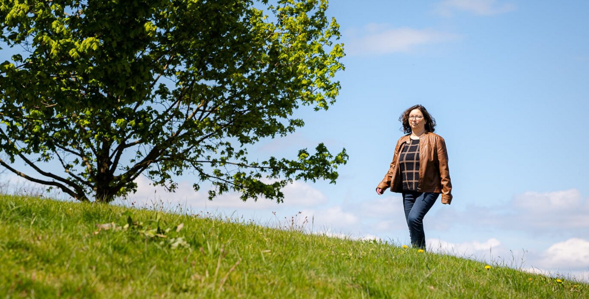 Katrin, een lymfoedeempatiënte, wandelt over een groene weide onder een heldere blauwe hemel. Op de achtergrond staat een grote, volle boom die een zomerse en rustige sfeer uitstraalt. Ze draagt een bruin leren jack en een spijkerbroek.