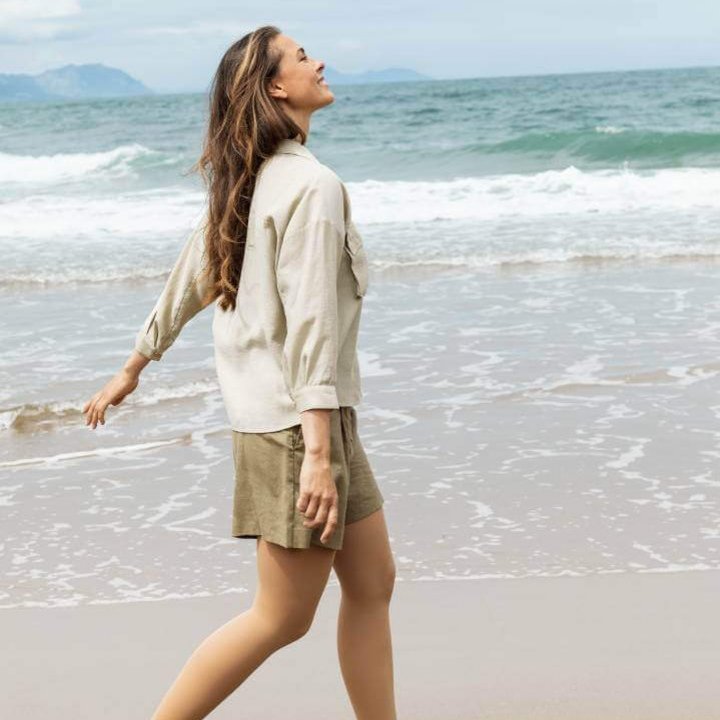 Een vrouw draagt VenoTrain soft compressiekousen en wandelt ontspannen langs het strand. Ze draagt een beige hemd, een korte olijfgroene broek en witte sneakers.